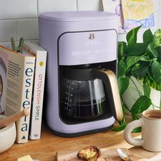 a purple coffee maker sitting on top of a wooden cutting board next to a book