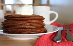 a stack of chocolate pancakes with whipped cream on top sitting on a white plate next to two coffee mugs