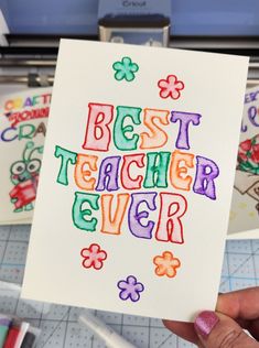 a person holding up a card with the words best teacher ever written in colorful letters