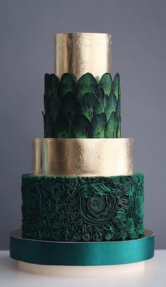 a three tiered green and gold wedding cake with flowers on the top, sitting on a table