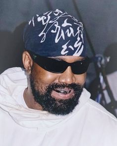 a man wearing sunglasses and a hat with writing on the side of his head, standing in front of a microphone