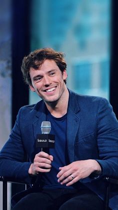 a man sitting in a chair holding a microphone and smiling at the camera while wearing a blue jacket