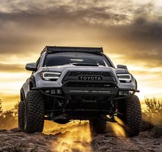 the front end of a gray toyota truck driving down a dirt road at sunset with clouds in the background