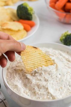 a hand holding a cracker over a bowl of dip with carrots and broccoli in the background