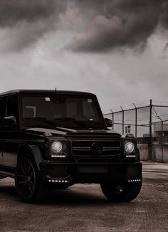 a black mercedes g class parked in front of a fenced off area with dark clouds