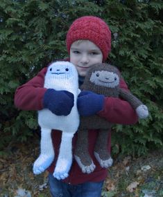 a young boy holding two stuffed animals in front of some bushes and trees with snow on the ground