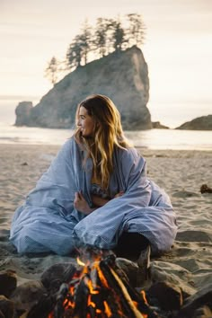 a woman wrapped in a blanket sitting next to a campfire on the beach at sunset