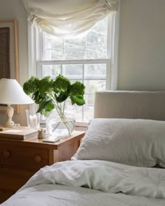 a bed with white sheets and pillows next to a lamp on a table in front of a window