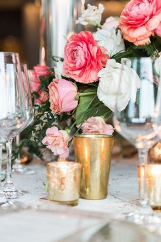 pink and white flowers in gold vases next to wine glasses on a table with candles