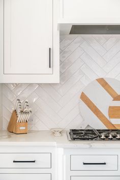 a kitchen with white cabinets and wooden cutting board on the stove top, next to utensils