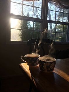 two coffee cups with steam rising from them on a table in front of a window