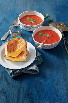 two bowls of tomato soup and grilled cheese sandwich on a blue tablecloth with spoons