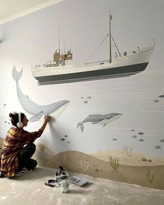 a woman is painting a wall with a boat and whale mural on the wall behind her