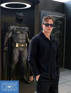 a man wearing sunglasses standing in front of a batman statue with his hands in his pockets