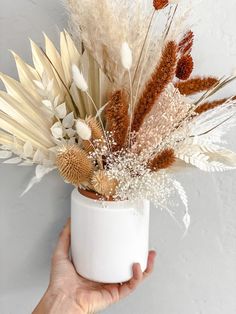 a hand holding a white vase with dried flowers in it and some feathers sticking out of the top