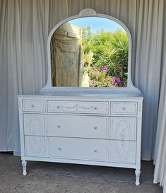 a white dresser with a large mirror on it's top and flowers in the background