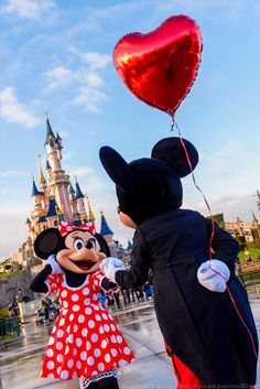 mickey and minnie mouse holding hands in front of a castle with a heart shaped balloon