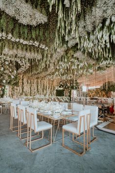 a long table with white chairs and flowers hanging from it's ceiling is surrounded by greenery