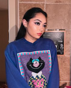 a woman with black eyeliners is wearing a blue sweatshirt that has an image of a cat on it