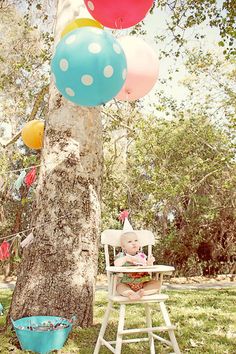 a baby sitting in a high chair next to a tree with balloons floating from it