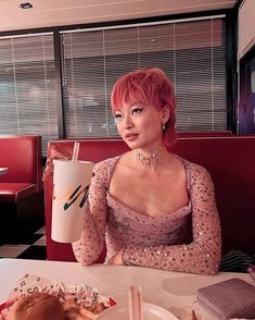 a woman with pink hair sitting at a table in front of a hamburger and drink