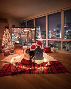 two people sitting on the floor in front of a christmas tree watching tv at night