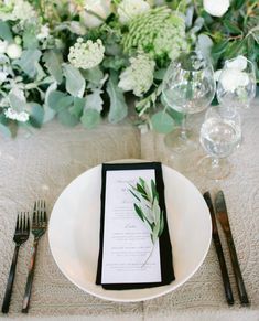 a place setting with white flowers and greenery on the table, along with silverware