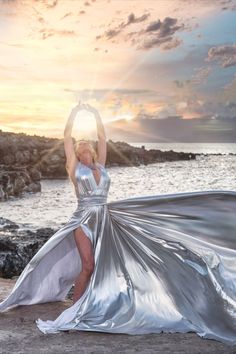 a woman in a silver dress is standing on the beach with her arms raised up