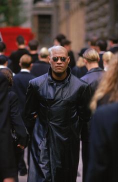 a man in a black coat and sunglasses walking down the street with other people behind him