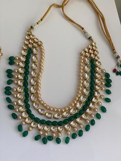 This luxurious necklace set epitomizes royal grandeur with its rich combination of green and gold. The set features: 1. **Necklace     - **Top Layer A row of pearly beads complemented by green beads, creating a harmonious blend of colors.    - **Middle Layer An alternating pattern of clear crystal settings and green beads, framed with gold accents, adding a touch of sophistication.    - **Bottom Layer A beautiful arrangement of pearls, green beads, and crystal settings, all culminating in a lavish design that radiates elegance. 2. **Earrings     - Matching earrings with a floral design composed of clear crystals and pearls, adorned with green bead drops, perfectly echoing the necklace's motif. The intricate craftsmanship and regal color scheme make this set an ideal choice for grand celebr Groom Jewelry, Royal Necklace, Luxurious Necklace, Earrings Matching, Necklace Top, 2 Earrings, Green Beads, Kundan Necklaces, Clear Crystals