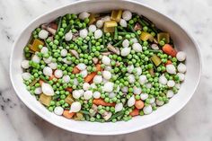 a white bowl filled with peas, carrots and other veggies on top of a marble counter