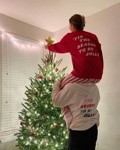 a woman in a red shirt is hanging on the back of a child's head next to a christmas tree