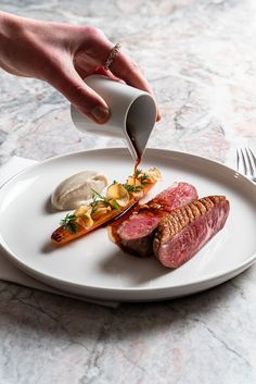 a person is pouring sauce on a plate with meat and potatoes in the background,