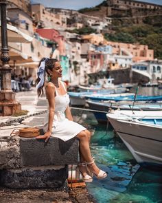 a woman sitting on a stone ledge next to boats in the water and buildings behind her