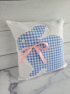 a blue and white gingham pillow with a pink bow on the front sitting on a marble counter