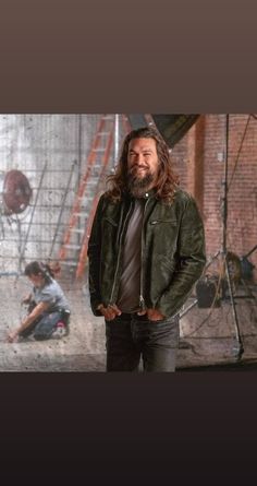 a man with long hair and beard standing in front of a brick wall wearing a green jacket