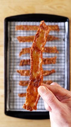 someone is holding up some bacon sticks in front of a pan with other food on it