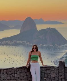 a woman standing on top of a stone wall next to the ocean with mountains in the background