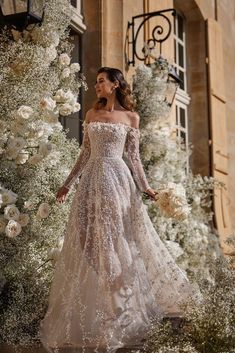 a woman in an off the shoulder wedding dress standing next to white flowers and greenery