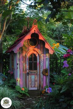 a small wooden house in the middle of some plants and trees with flowers around it