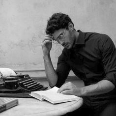 a man sitting at a desk in front of an old typewriter and reading a book