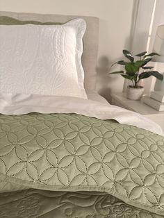 a white bed with green comforter and pillows on it's headboard, next to a potted plant