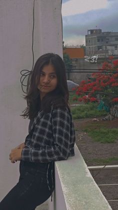 a woman leaning against a white wall with red flowers in the back ground and buildings in the background