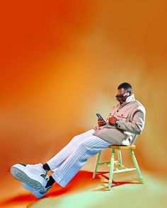 a man sitting on a stool using a cell phone while wearing white pants and a tan jacket