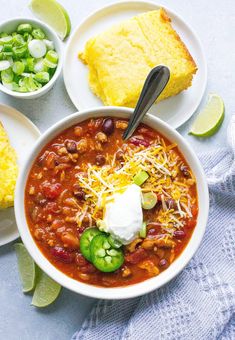 two bowls of chili with sour cream, cornbreads and green onions on the side