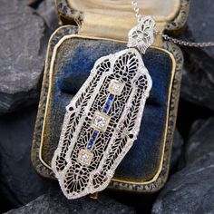 This breathtaking circa 1930s 14K white gold filigree pendant features engraved details and is bordered with milgrain edging. The pendant is accented with three (3) bead set round transitional brilliant cut diamonds, and eight (8) channel set square French cut lab grown sapphires. The pendant measures 53.3mm in length (including bail) by 20.0mm in width. The pendant is suspended from a platinum cable link neck chain measuring 16 inches in length. Antique White Gold Filigree Necklace, Vintage White Gold Necklace With Diamond Accents, Antique White Gold Necklace With Filigree, Antique White Gold Necklace With Intricate Design, Formal White Gold Filigree Jewelry, Art Deco Diamond Necklace With Filigree Details, Art Deco Diamond Necklace With Filigree, Art Deco White Gold Jewelry With 17 Jewels, Classic Pendant Necklace With Filigree