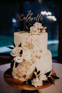 a white wedding cake with gold decorations and flowers on the top is sitting on a wooden stand