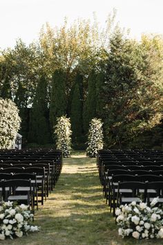 an outdoor ceremony setup with black chairs and white flowers on the aisle, surrounded by trees