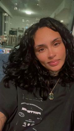 a woman with long black curly hair wearing a t - shirt and looking at the camera