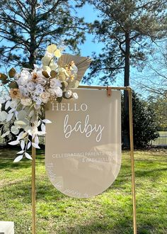 a hello baby sign with flowers on it in front of some grass and trees at a park